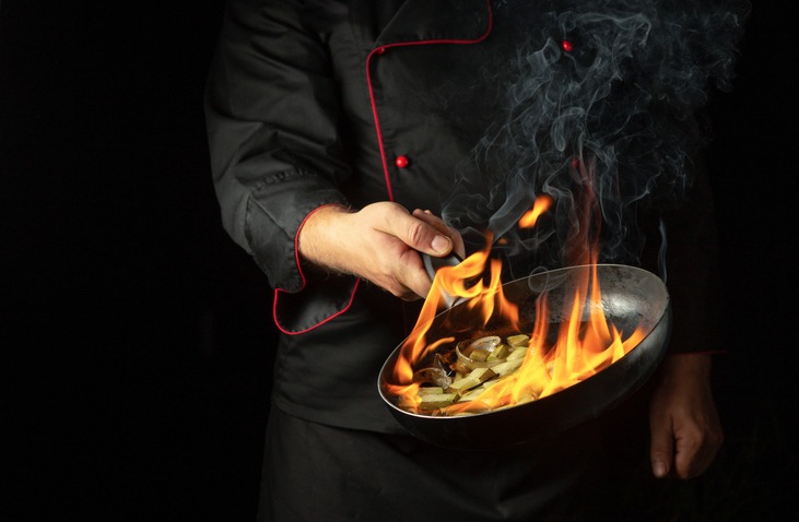 a chef cooking food on a frying pan with flame of fire from rum